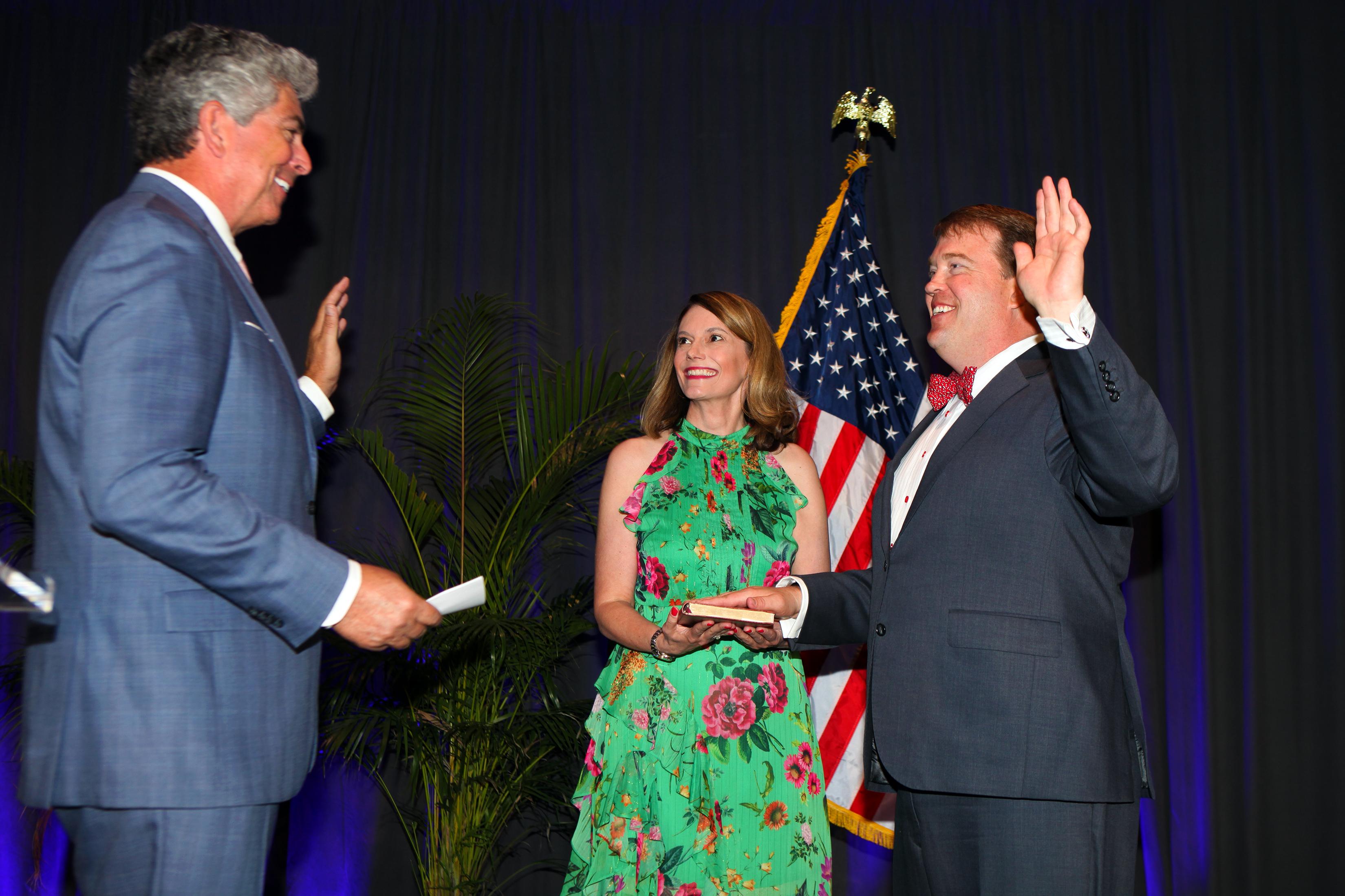 Man in gray suit with right hand raised standing next to a woman in a green dress holding a Bible, and a tall man with gray hair in a gray suit administering an oath